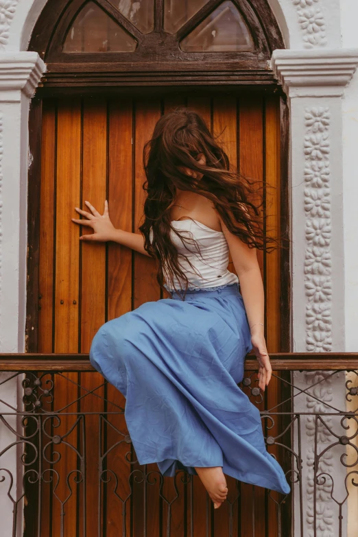 a woman standing on a balcony next to a wooden door, a picture, by Alejandro Obregón, pexels contest winner, arabesque, playful pose, her loose hair, a blue skirt, 15081959 21121991 01012000 4k
