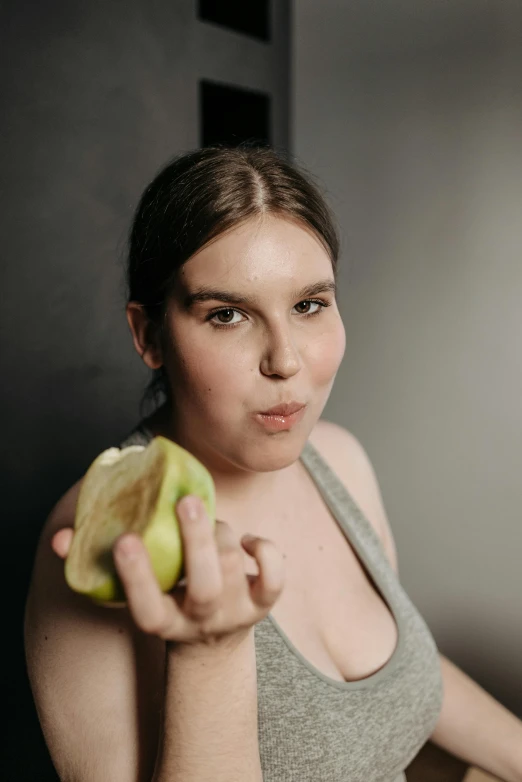 a woman holding an apple in front of her face, an album cover, inspired by Artemisia Gentileschi, trending on pexels, patricia piccinini, she is wearing a black tank top, having a snack, lime