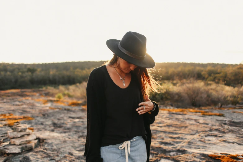 a woman standing on top of a rocky field, pexels contest winner, minimalism, black stetson hat, wearing in cardigan, in the australian outback, bracelets and necklaces