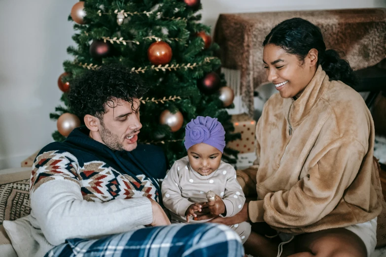 a man and woman sitting on a couch with a baby in front of a christmas tree, a photo, pexels, incoherents, mixed race, manuka, portrait image