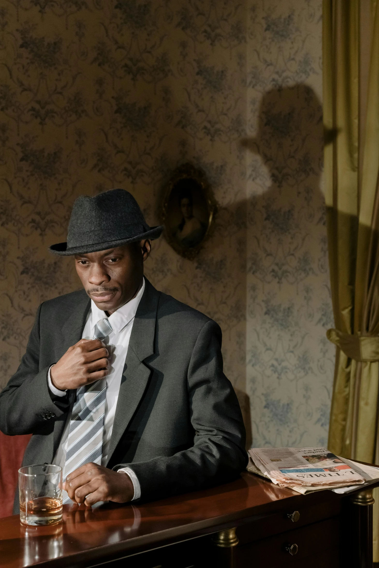 a man in a suit and hat sitting at a table, an album cover, unsplash, harlem renaissance, ( ( theatrical ) ), model pose, thought provoking, backdrop