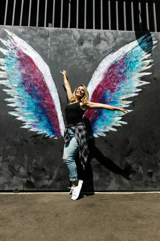 a woman standing in front of a wall with wings painted on it, chalk art, by Arabella Rankin, pexels contest winner, brandi love, triumphant pose, tourist photo, evanna lynch