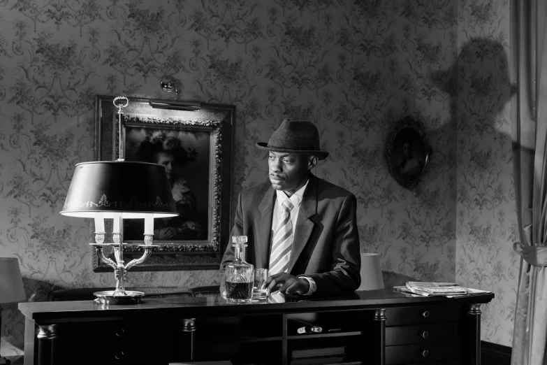 a black and white photo of a man sitting at a desk, by Maurycy Gottlieb, pexels, harlem renaissance, a suited man in a hat, scene from a 1 9 7 3 film, tyler the creator, standing in a dimly lit room