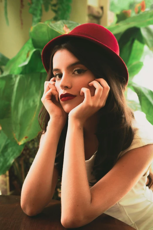 a woman sitting at a table talking on a cell phone, an album cover, inspired by Elsa Bleda, instagram, renaissance, maroon hat, isabela moner, headshot photo, plants