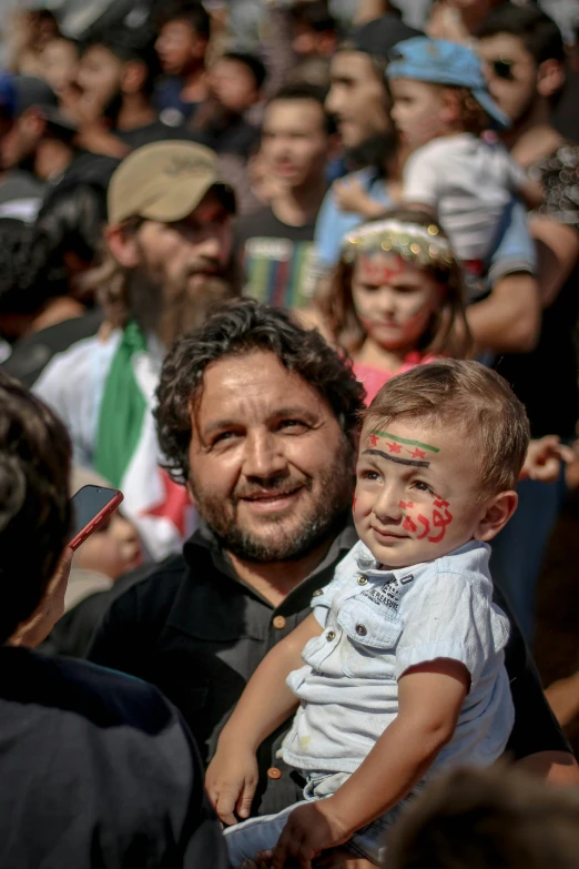 a man holding a child in front of a crowd of people, arab man light beard, markings on his face, promo image, color photograph