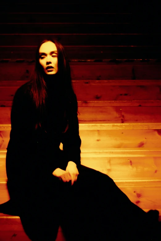 a woman sitting on top of a wooden bench, an album cover, international gothic, standing in a dimly lit room, with long black hair, pale complexion, yohji yamamoto