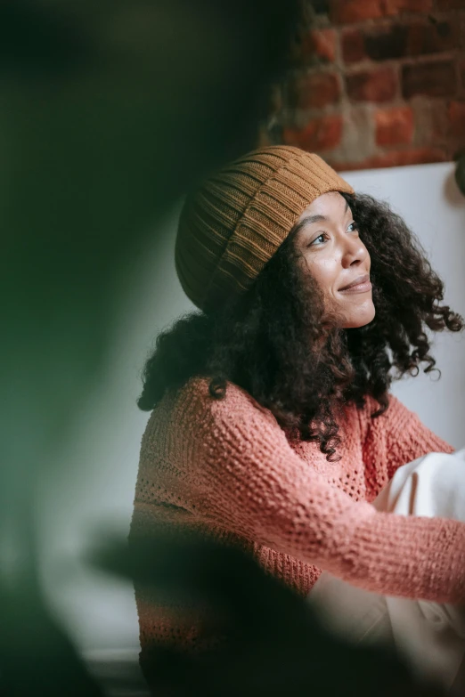 a woman sitting in front of a brick wall, trending on pexels, renaissance, knitted hat, mixed-race woman, sitting across the room, wearing a red turtleneck sweater