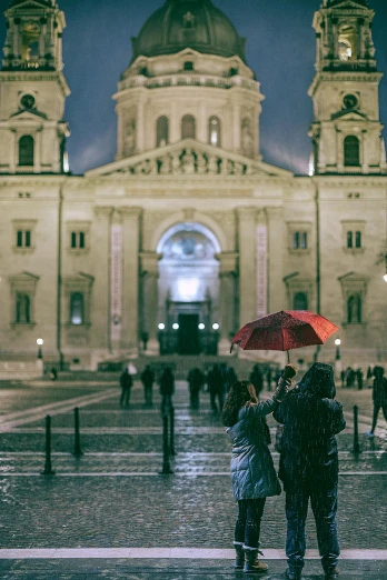 a group of people standing in front of a large building, by Adam Szentpétery, unsplash contest winner, rainy night, budapest, cathedral, couple kissing