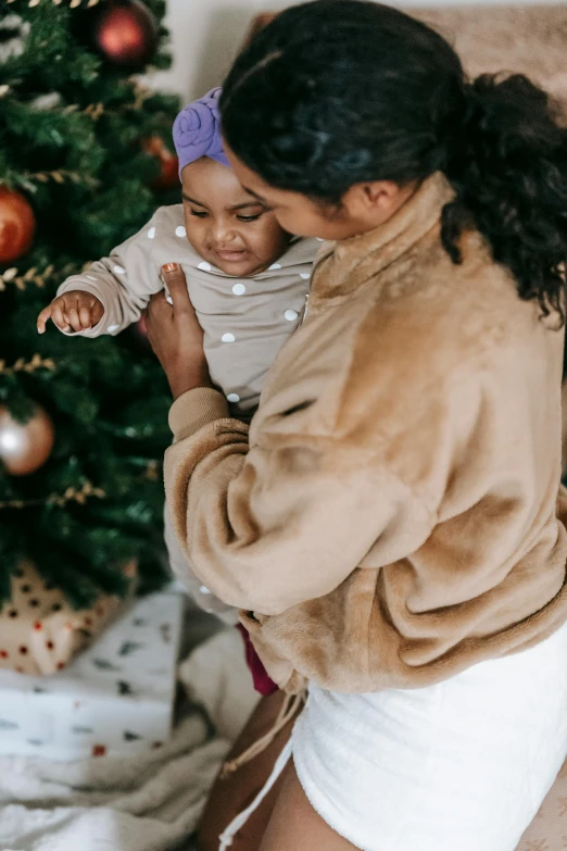 a woman holding a baby in front of a christmas tree, pexels, tan and brown fur, manuka, brown clothes, thumbnail