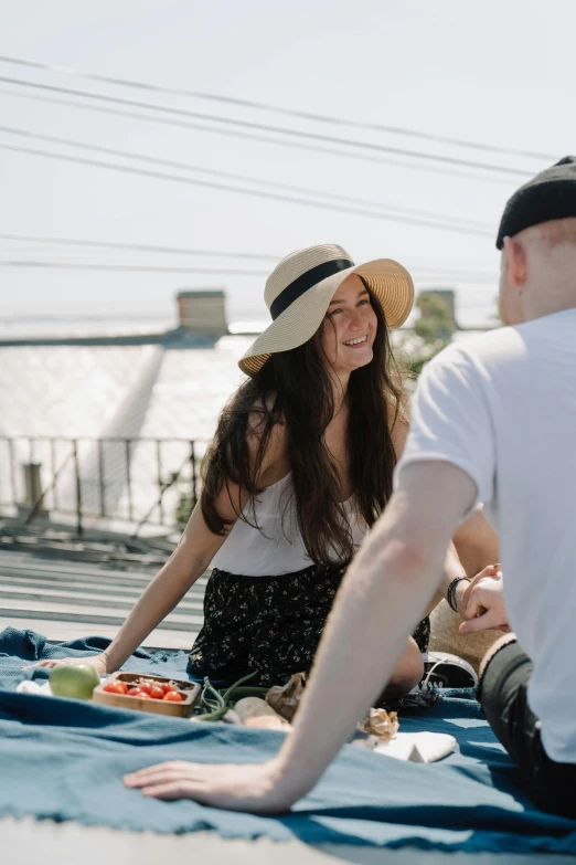 a group of people sitting on top of a blue blanket, wearing a hat, food, on a bridge, smiling couple