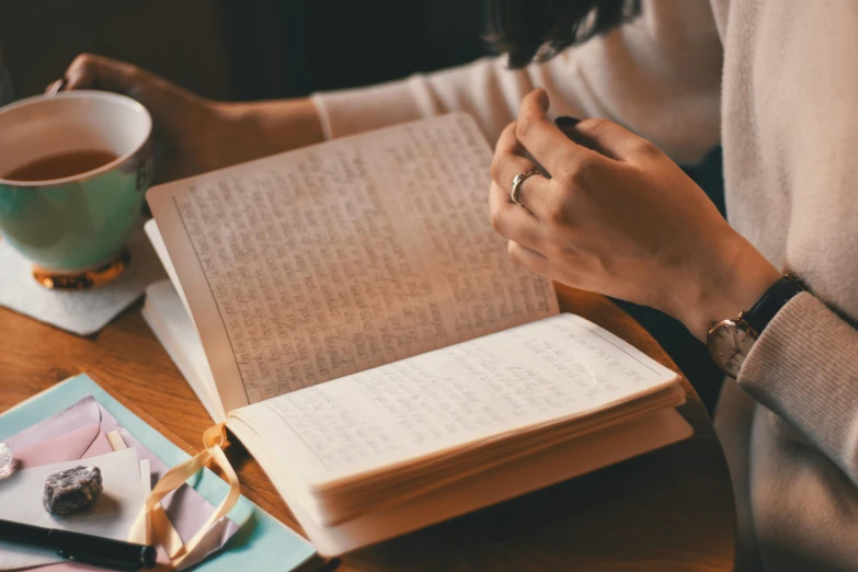 a woman sitting at a table with a book and a cup of coffee, pexels contest winner, private press, holding notebook, old books and dip pen, opening shot, found written in a notebook