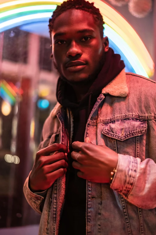 a man standing in front of a neon sign, by Cosmo Alexander, trending on pexels, realism, brown jacket, non binary model, ( ( dark skin ) ), jean jacket