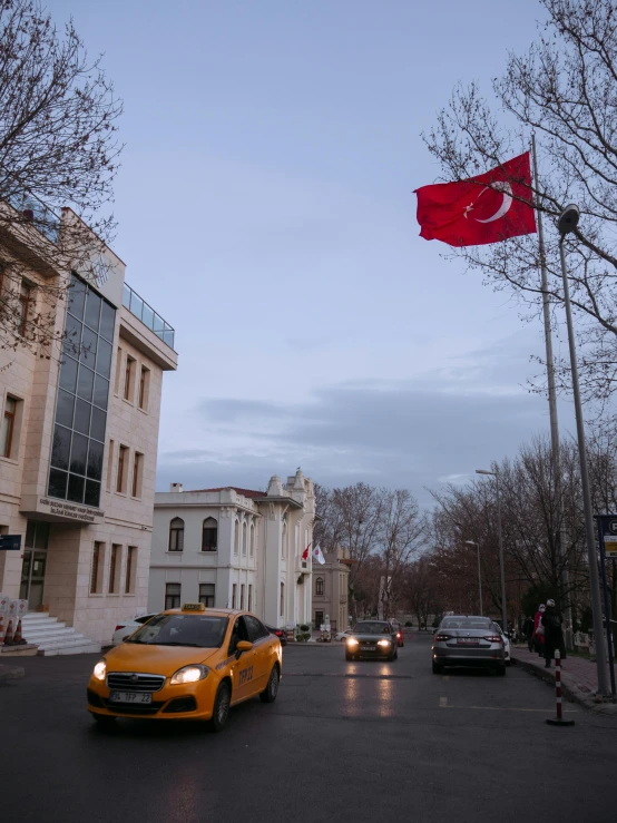 a yellow car driving down a street next to a tall building, an album cover, inspired by Niyazi Selimoglu, pexels contest winner, hurufiyya, flag, turkey, today\'s featured photograph 4k, late evening