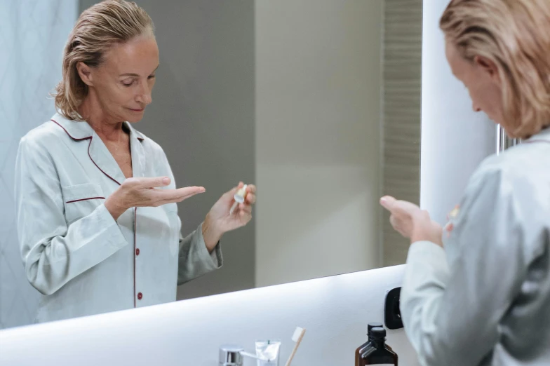 a woman that is standing in front of a mirror, quack medicine, hands on counter, profile image, wearing a grey robe