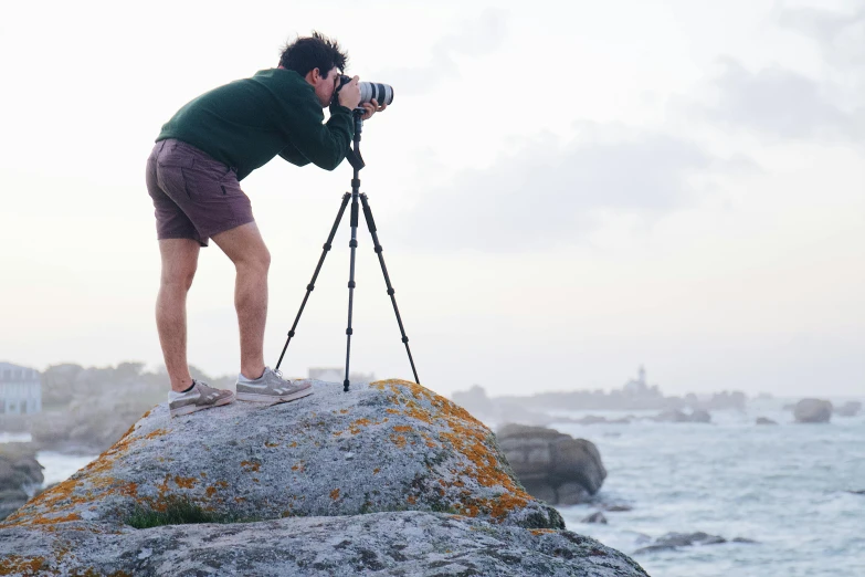 a man standing on top of a rock next to the ocean, by Carey Morris, pexels contest winner, with nikon cameras, tripod, standing on boulder, camera close to the legs