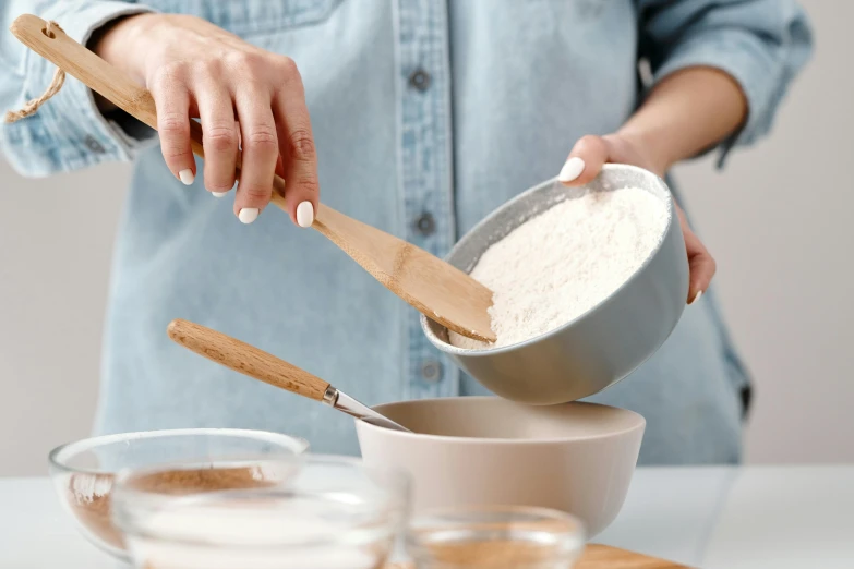 a woman mixing ingredients in a bowl on a table, trending on pexels, grey, background image, bakery, spatula