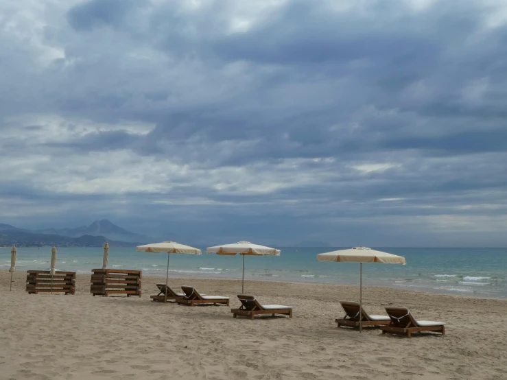 a group of lounge chairs sitting on top of a sandy beach, grey cloudy skies, greek setting, profile image, umbrellas