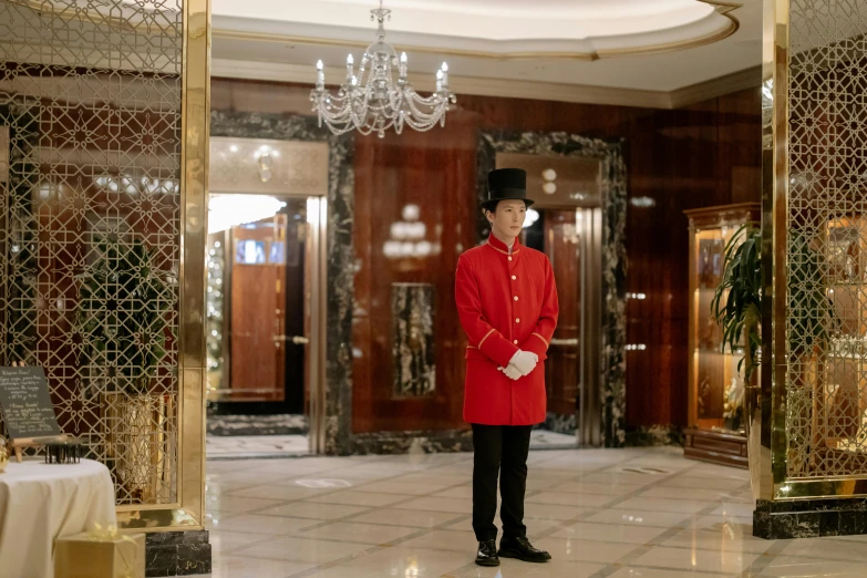 a man in a red uniform standing in front of a chandelier, hotel, full body black and red longcoat, red coat, photographed for reuters