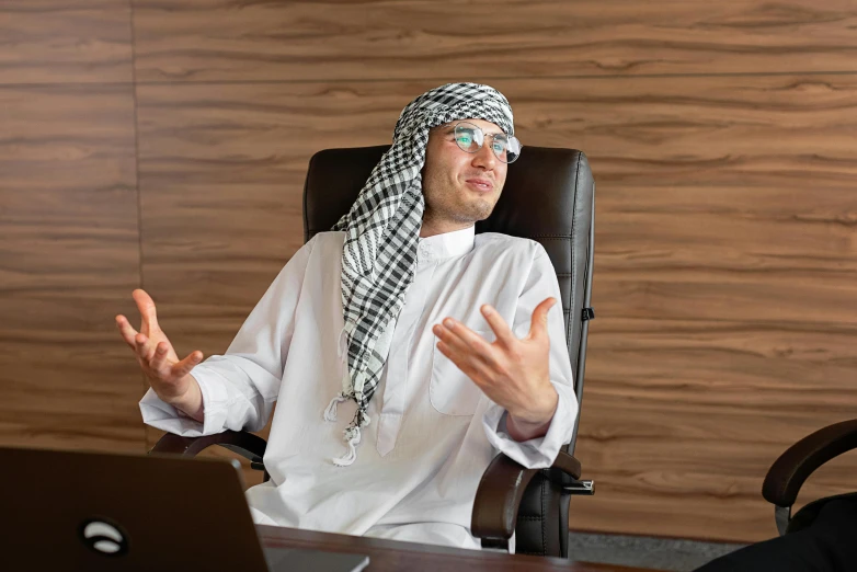 a man sitting in a chair in front of a laptop, inspired by Sheikh Hamdullah, pexels, hurufiyya, shrugging arms, funny professional photo, traditional clothing, typical cryptocurrency nerd