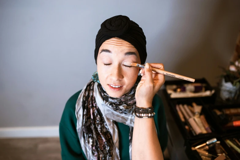 a woman is putting makeup on her face, inspired by Ina Wong, hurufiyya, wearing a head scarf, smooth brushwork, complimentary eyeliner, looking down on the camera