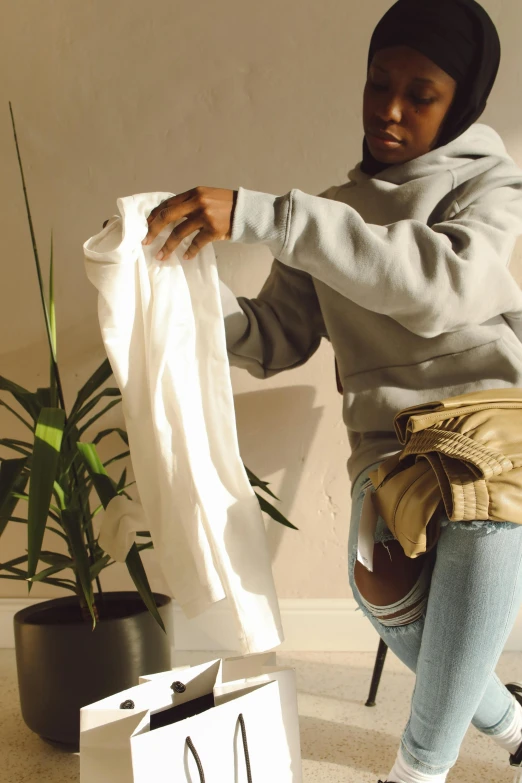 a woman sitting on a chair holding a bag, by Nina Hamnett, happening, beige hoodie, houseplant, double layer fold over hem, handsome hip hop young black man