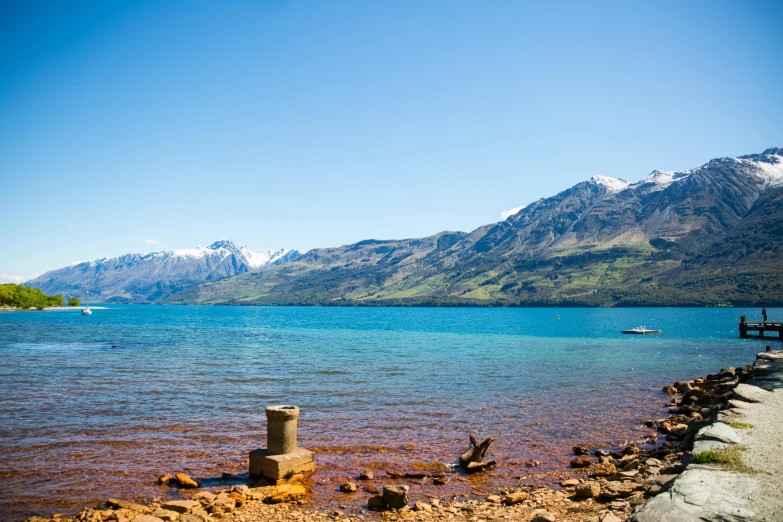 a body of water with mountains in the background, by Julia Pishtar, unsplash, hurufiyya, manuka, clear blue skies, on a pedestal, shore of the lake