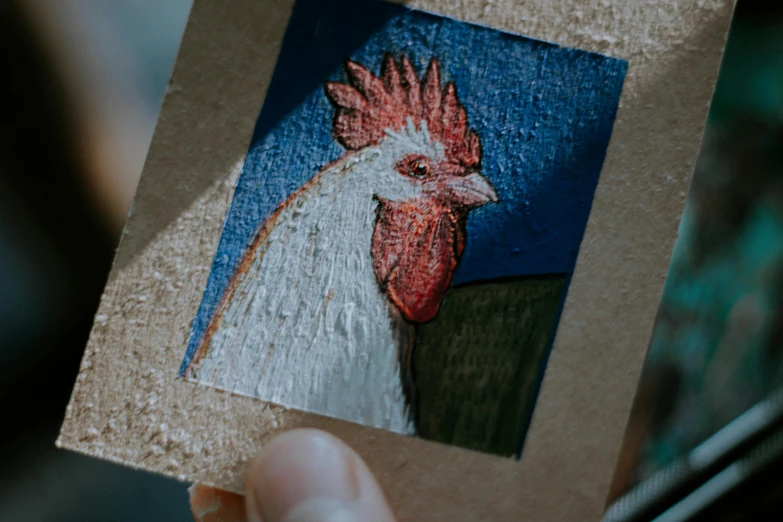 a close up of a person holding a picture of a rooster, by Julia Pishtar, arbeitsrat für kunst, cardboard, enamel, detailed hatching, trading card