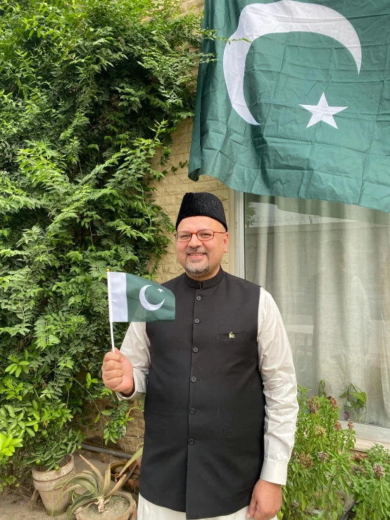 a man standing in front of a house holding a pakistan flag, an album cover, pexels contest winner, hurufiyya, taken on iphone 14 pro, 2 5 6 x 2 5 6 pixels, 0, posing