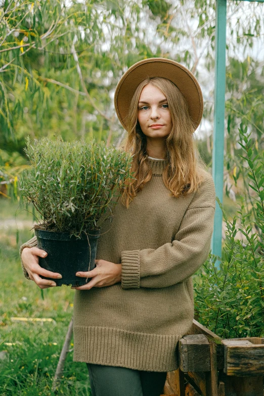 a woman in a hat holding a potted plant, a portrait, trending on pexels, wearing an oversized sweater, manuka, with a garden, handsome girl