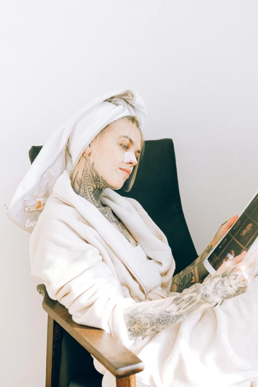 a woman sitting in a chair reading a book, an album cover, inspired by Louisa Matthíasdóttir, trending on pexels, facial tattoos, bathrobe, covered head, skincare