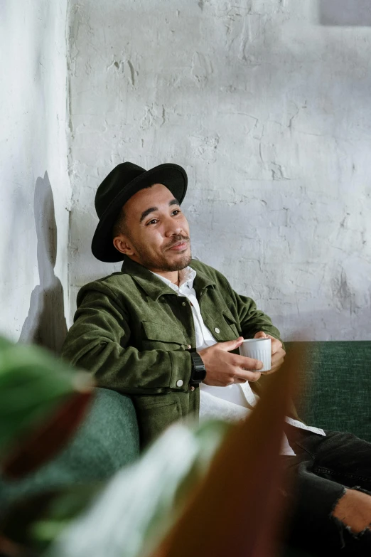 a man sitting on a couch with a cup of coffee, a portrait, pexels contest winner, green hat, robert sheehan, singer maluma, andrew thomas huang