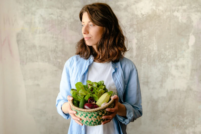 a woman holding a basket full of vegetables, by Nicolette Macnamara, pexels contest winner, greens and blues, on grey background, salad, portrait of small