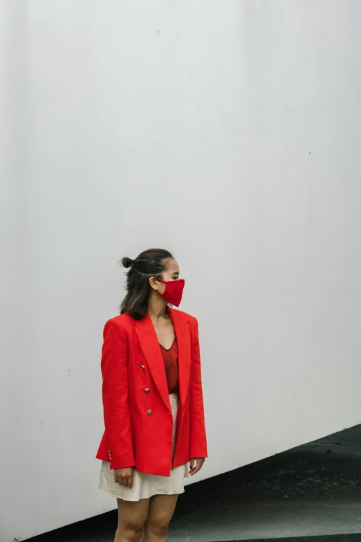 a woman standing in front of a white wall, by Olivia Peguero, pexels contest winner, villain wearing a red oni mask, trench coat and suit, gemma chen, wearing facemask