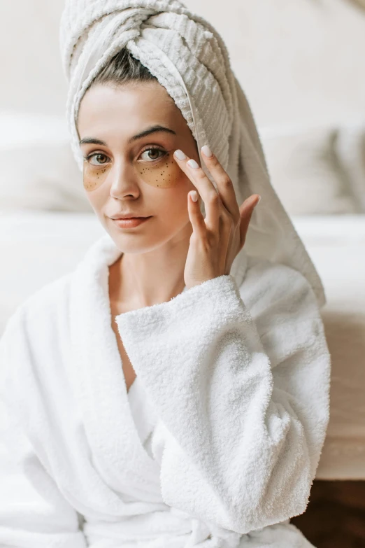 a woman sitting on a bed with a towel on her head, by Julia Pishtar, trending on pexels, renaissance, dark circles under bemused eyes, gold and white robes, wearing small round glasses, skincare