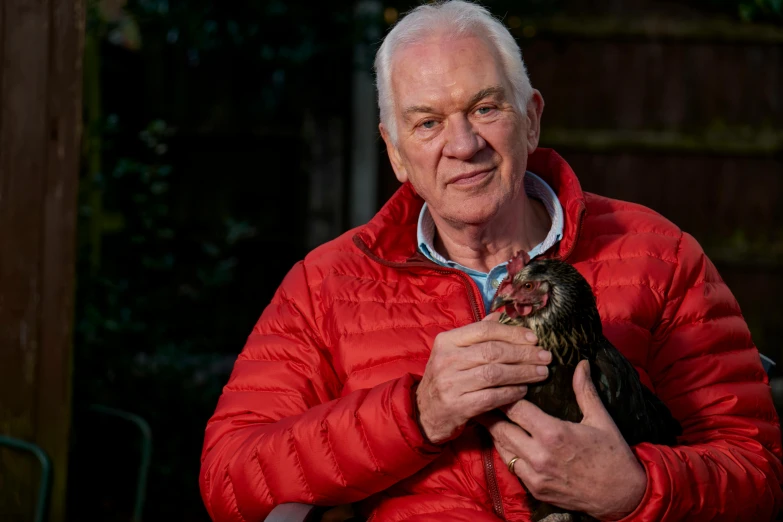 a man in a red jacket holding a chicken, a portrait, by Paul Bird, close to the camera, portrait image, an elderly, thumbnail