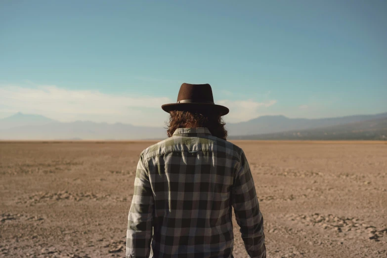 a man standing in the middle of a desert, an album cover, unsplash, caracter with brown hat, heath clifford, back facing the camera, unkept hair