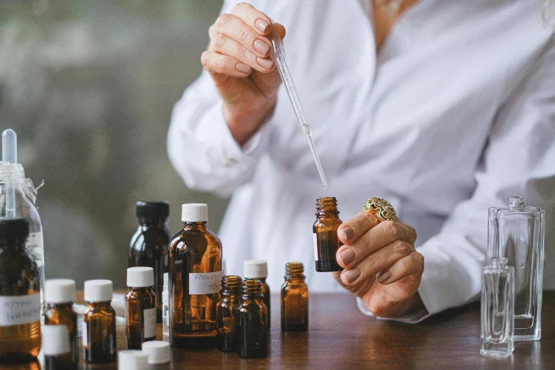 a woman sitting at a table with bottles of essential oils, by Daniel Lieske, trending on pexels, with a lab coat, wearing brown robes, manuka, hemp