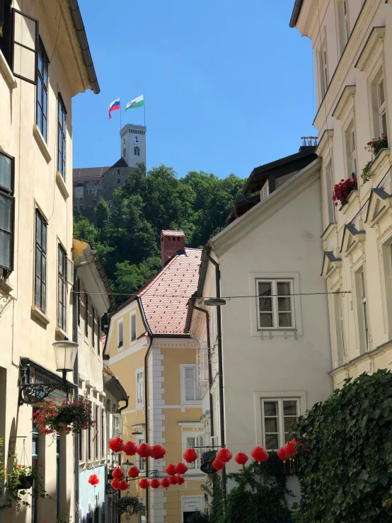 a narrow street with a clock tower in the background, by Emma Andijewska, castle on the mountain, square, wires hanging above street, red peaks in the background