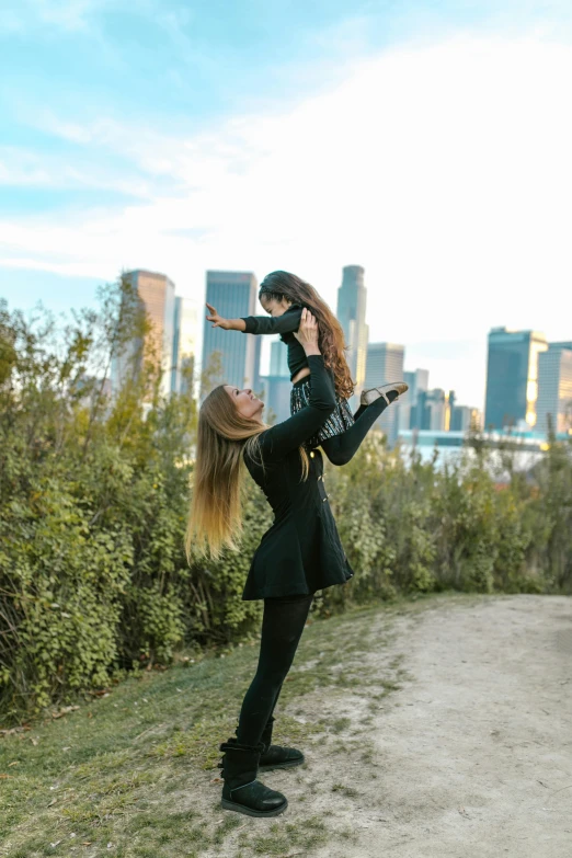 a woman standing on top of a dirt road, with a kid, perched on a skyscraper, ballet style pose, at a park