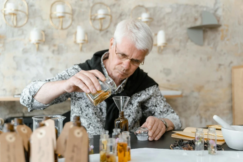 a man sitting at a table pouring something into a glass, pexels contest winner, local conspirologist, alembic, older male, artisan