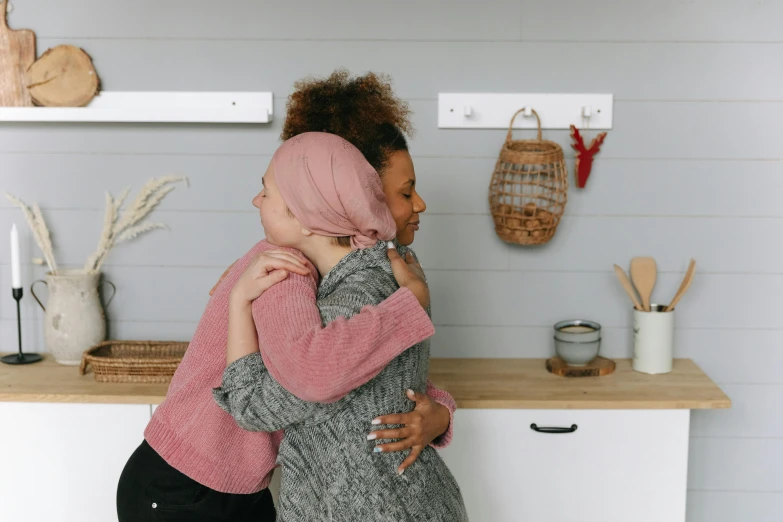 two women hugging each other in a kitchen, pexels contest winner, hurufiyya, pink and grey muted colors, varying ethnicities, no duplicate image, background image