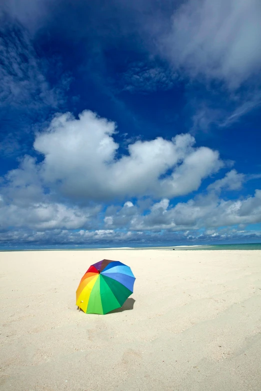 a colorful umbrella sitting on top of a sandy beach, by Peter Churcher, white sand beach, cumulus, square, rainbow