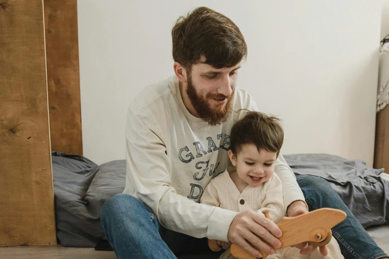a man sitting next to a little boy on a bed, pexels contest winner, wooden art toys, avatar image, small beard, maintenance photo
