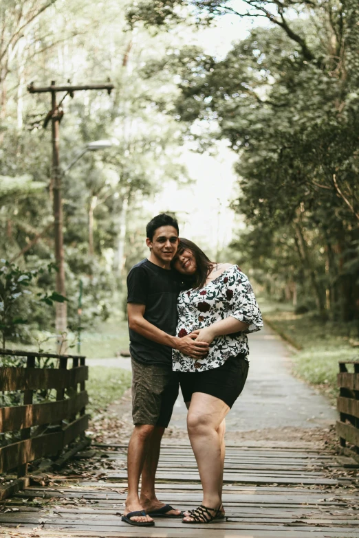 a man and a woman standing on a wooden bridge, lush surroundings, pregnant, in sao paulo, hugging