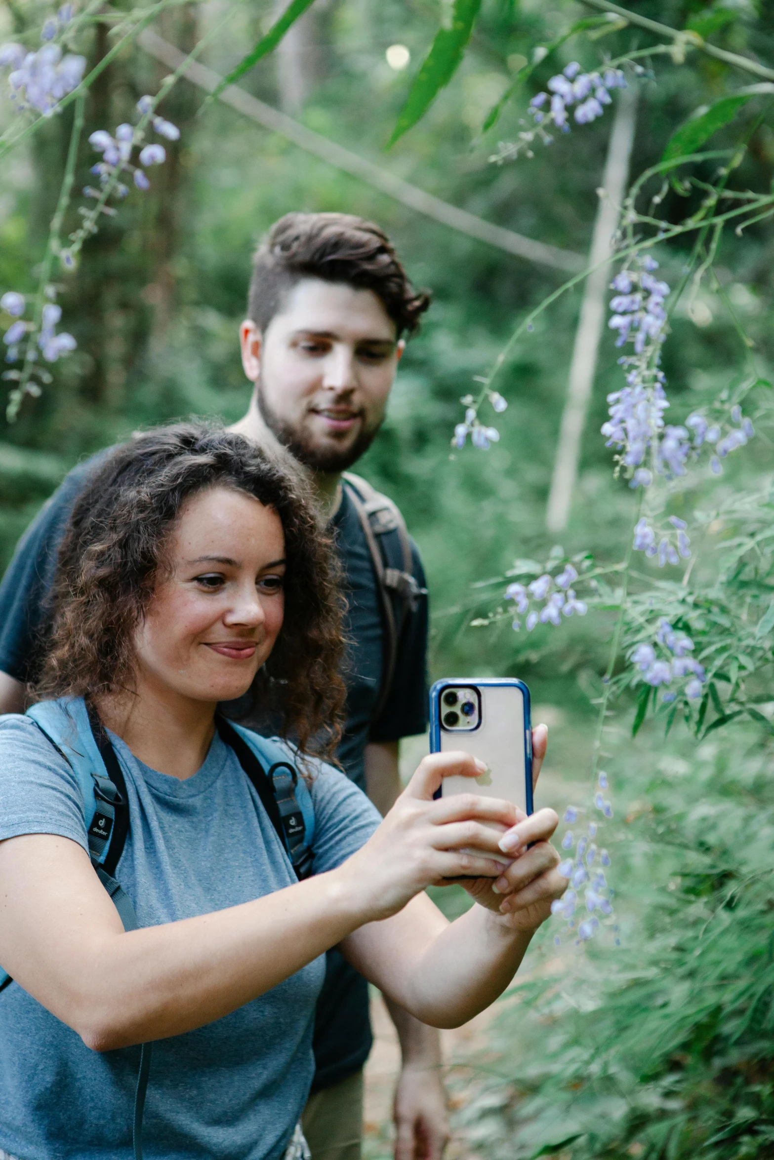 a man and woman taking a selfie in the woods, flowers around, humans exploring, casually dressed, phone