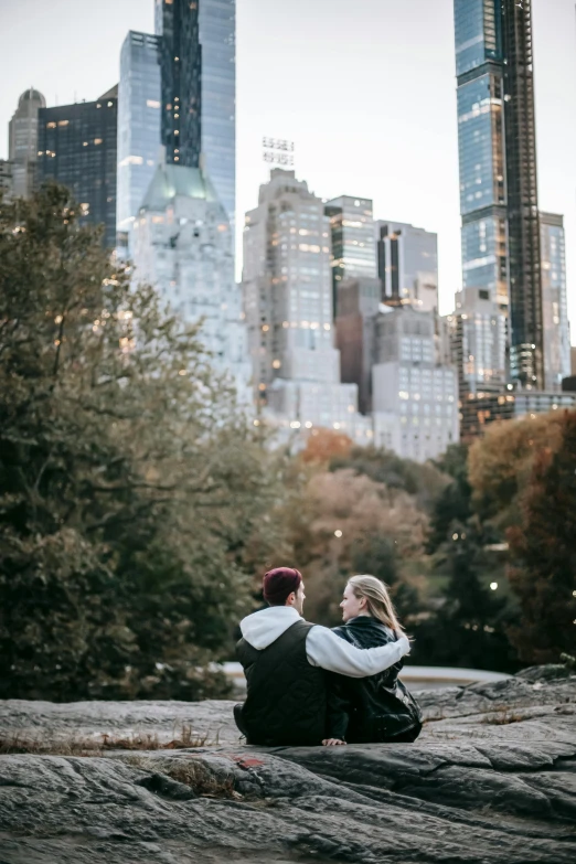 a couple of people sitting on top of a rock, in new york, trees in the background, hugging, profile pic