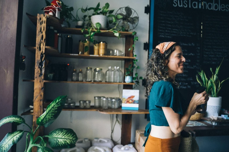 a woman standing in front of a shelf filled with potted plants, by Julia Pishtar, trending on unsplash, happening, aussie baristas, holding a baguette, a person standing in front of a, ariel perez