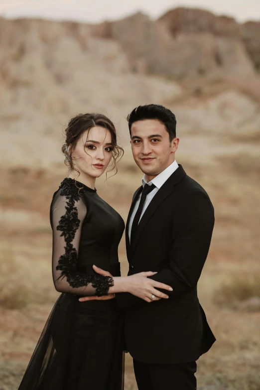 a man in a tuxedo standing next to a woman in a black dress, a portrait, by irakli nadar, pexels contest winner, hills in the background, square, pale-skinned persian girl, modeled