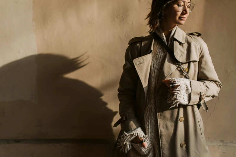 a woman in a trench coat standing next to a wall, by Nina Hamnett, trending on pexels, gloves and jewelry. motion, with sunglass, nishihara isao, feathers growing from arms