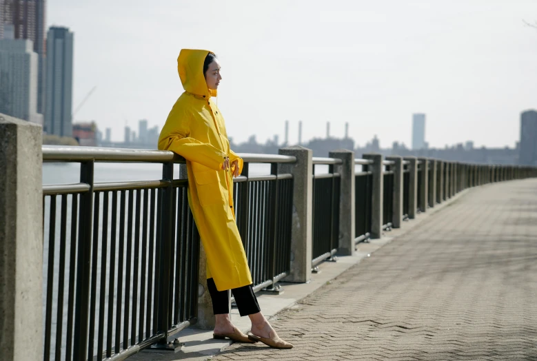 a woman in a yellow raincoat leaning against a fence, by Nina Hamnett, unsplash, at the waterside, brooklyn, wearing nanotech honeycomb robe, full body wide shot